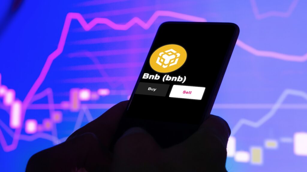 Smartphone screen displaying the Binance Coin (BNB) logo with Buy and Sell buttons, held against a neon blue stock-chart background.