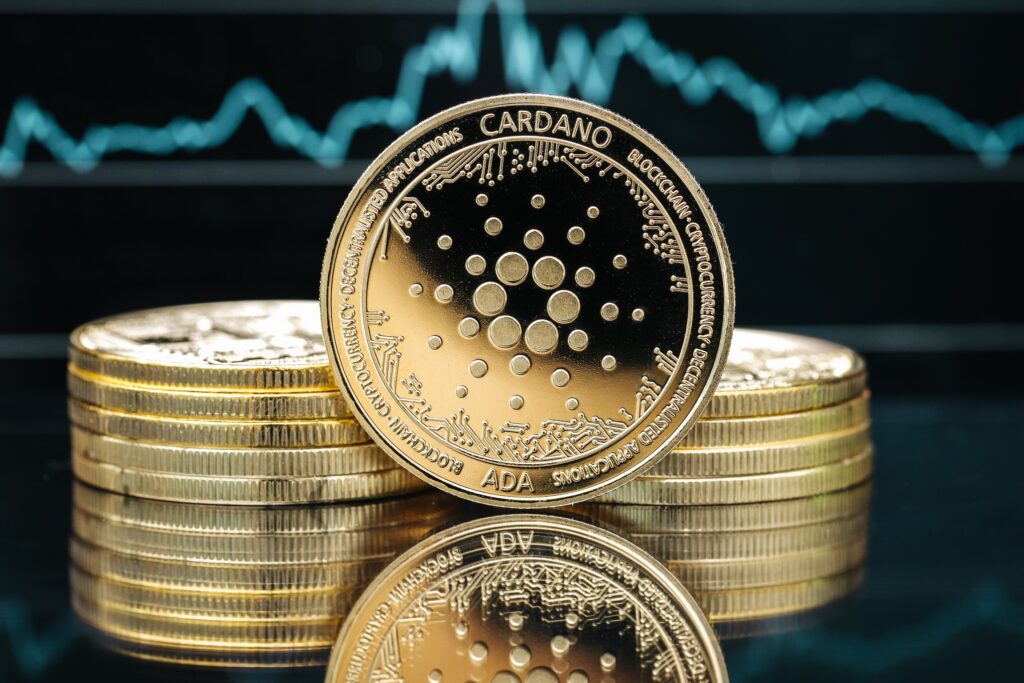 Close-up of Cardano ADA coins stacked on a reflective surface with a blue stock/price chart in the background