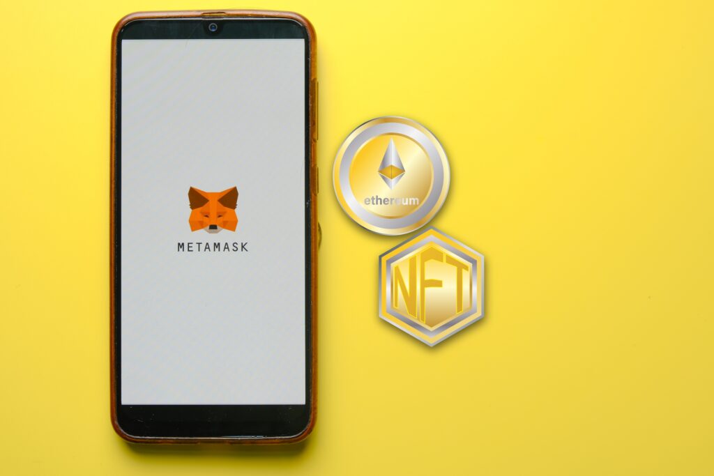 Smartphone screen shows the MetaMask logo with crypto icons (Ethereum coin and NFT badge) on a bright yellow background.