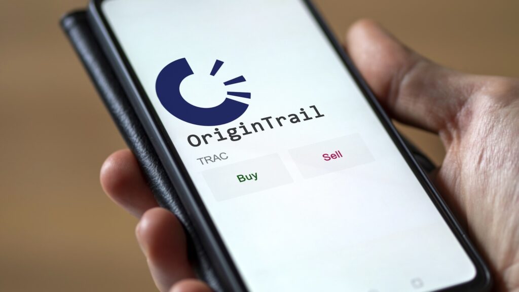 OriginTrail - Crypto-Globe.com OriginTrail app on a smartphone screen showing a blue logo and Buy (green) and Sell (pink) buttons.
